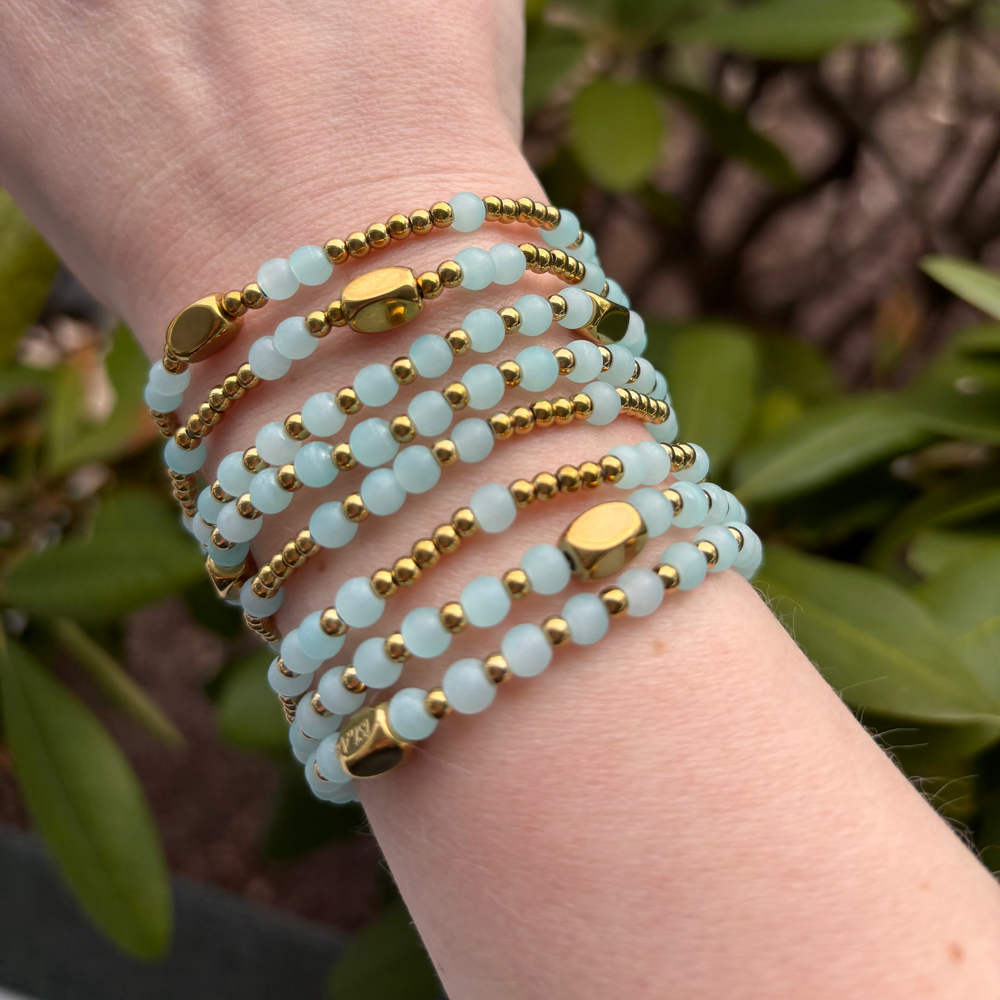 Hand wearing multiple light blue and gold beaded bracelets with a green leafy background
