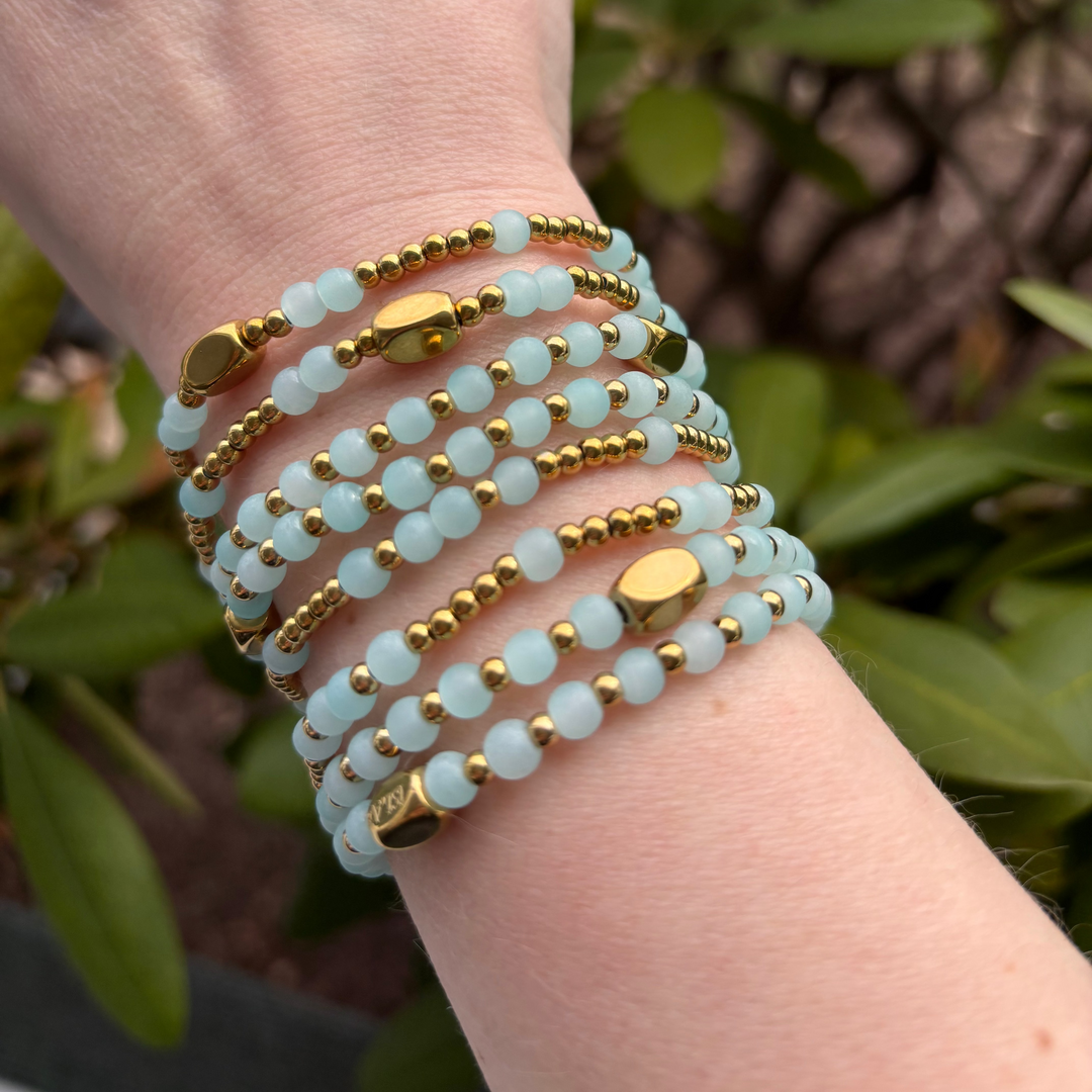 Hand wearing multiple light blue and gold beaded bracelets with a green leafy background
